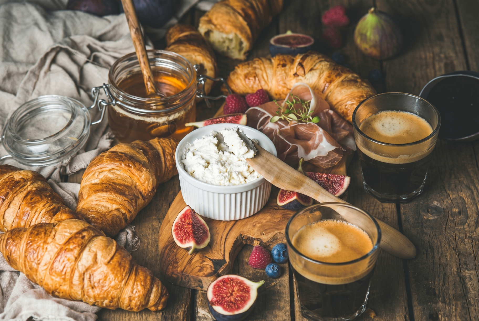 Rustic breakfast spread with open honey jar, fresh croissants, ricotta, figs, berries and espresso on a weathered wooden table